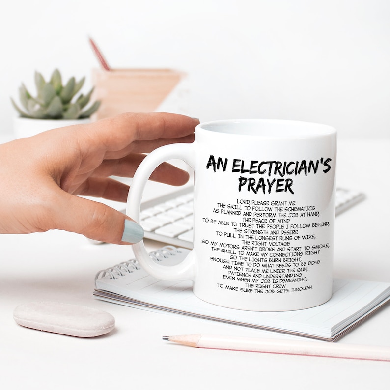 May include: White ceramic mug with the text "An Electrician's Prayer" in black. The mug features a list of aspirations for an electrician. The mug is held by a person with light blue nail polish.