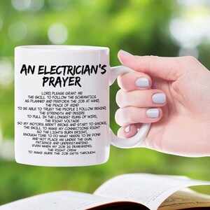 May include: White ceramic mug with the text "AN ELECTRICIAN'S PRAYER" in black. The mug features a prayer for electricians. The mug is held by a person with a blurred green background.