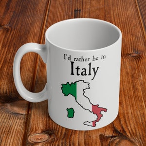 Puede incluir: Taza de cer&aacute;mica blanca con un contorno negro de la pen&iacute;nsula italiana y los colores de la bandera italiana. El texto "Prefiero estar en Italia" est&aacute; impreso en la taza.