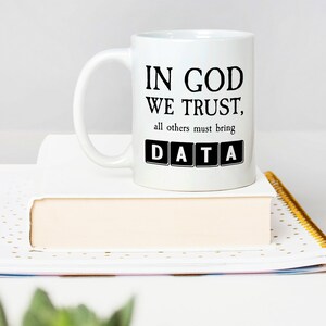 Op de afbeelding: Witte keramische mok met zwarte tekst die "In God We Trust, all others must bring DATA" luidt. Het woord DATA staat in zwarte vierkanten.