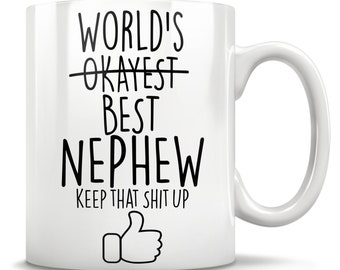 May include: White ceramic mug with black text: "WORLD'S BEST NEPHEW", with "OKAYEST" crossed out. Below is "KEEP THAT SHIT UP" and a thumbs-up graphic. The mug has a curved handle, ideal for tea or coffee.