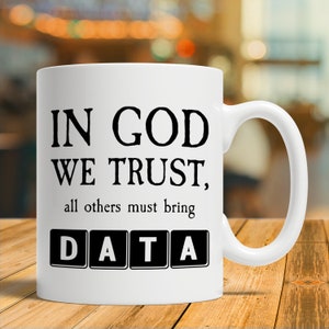 Op de afbeelding: Witte keramische koffiemok met zwarte tekst die "In God We Trust, all others must bring DATA" luidt. Het woord DATA staat in zwarte blokletters.