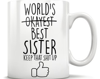 Puede incluir: Taza de café de cerámica blanca con texto negro que dice "World's Okayest Best Sister Keep That Shit Up" con un icono de pulgar hacia arriba.