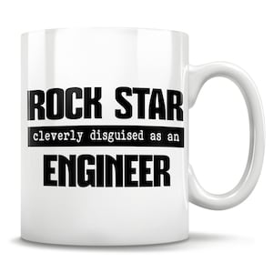 Könnte beinhalten: Weiße Keramik-Kaffeetasse mit schwarzem Text, der "Rock Star cleverly disguised as an Engineer" lautet.