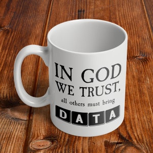 Op de afbeelding: Witte keramische mok met zwarte tekst die "In God We Trust, all others must bring DATA" luidt. Het woord DATA staat in zwarte blokletters.