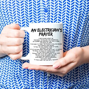 May include: White ceramic mug with the text "AN ELECTRICIAN'S PRAYER" in black. The mug features a prayer for electricians, with text in a black, easy-to-read font. The mug is held by a person wearing a blue and white patterned shirt.