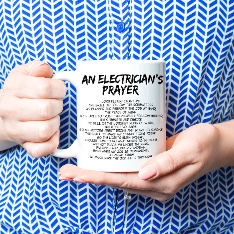 May include: White ceramic mug with the text "AN ELECTRICIAN'S PRAYER" in black. The mug features a prayer for electricians, with text in a black, easy-to-read font. The mug is held by a person wearing a blue and white patterned shirt.