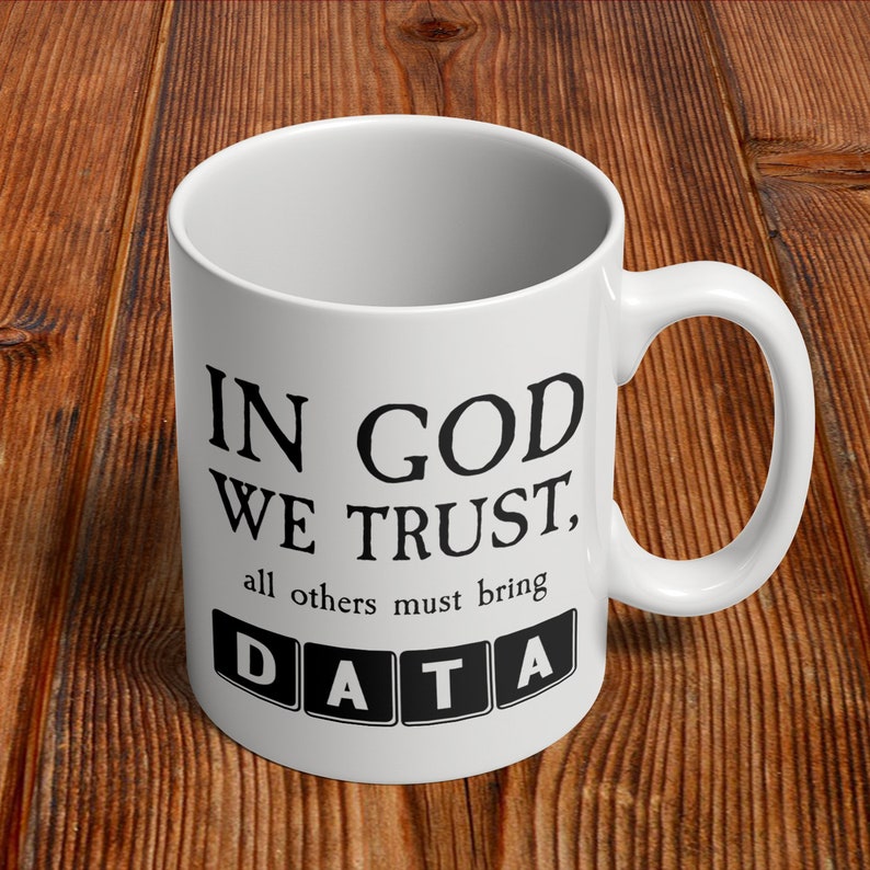 Op de afbeelding: Witte keramische mok met een zwarte inscriptie die luidt "In God We Trust, all others must bring DATA". Het woord DATA wordt weergegeven in zwarte vierkanten.