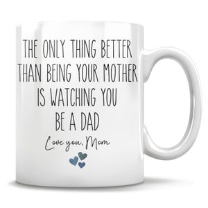 Op de afbeelding: Een witte keramische mok met zwarte bedrukte tekst die luidt: THE ONLY THING BETTER THAN BEING YOUR MOTHER IS WATCHING YOU BE A DAD. Onder de tekst staat Love you, Mom, gevolgd door drie kleine, blauwachtige hartpictogrammen onderaan.