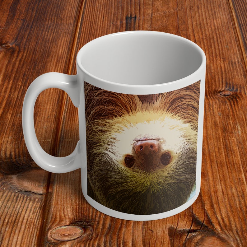 May include: White ceramic mug with a brown and tan sloth face printed on it. The sloth is upside down and looking directly at the viewer.