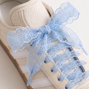 Op de afbeelding: Een crèmekleurige leren sneaker met drie witte zijstrepen en een rubberen zool. De schoen is geregen met een lichtblauw kanten lint dat bovenaan tot een strik is geknoopt, waardoor de standaard veters zijn vervangen door een decoratieve afwerking.