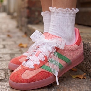 Op de afbeelding: Een paar roze en rode sneakers met textuur, groene zijstrepen, een doorschijnende roze zool en witte kanten strikken. De sneakers worden gedragen met witte enkelsokken die aan de bovenkant zijn afgewerkt met een fijne kanten rand.