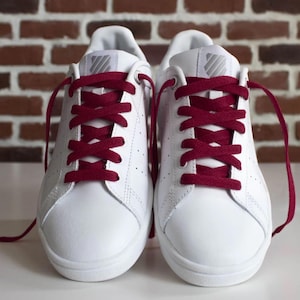 Può includere: Un paio di scarpe da ginnastica in pelle bianca con lacci bordeaux. Le scarpe hanno un design classico con un profilo basso e una punta traforata. I lacci sono ben legati, su sfondo di mattoni.