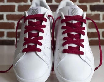 May include: A pair of white leather sneakers with burgundy laces. The shoes have a classic design with a low profile and a perforated toe box. The laces are neatly tied, and the shoes are set against a brick background.