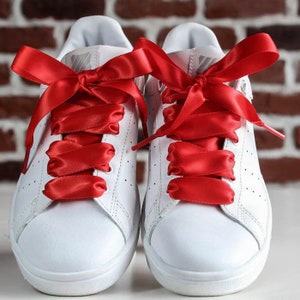 Può includere: Un paio di scarpe da ginnastica bianche con lacci in nastro di raso rosso brillante. I lacci sono annodati con un fiocco in alto. Le scarpe sono su uno sfondo di muro di mattoni.