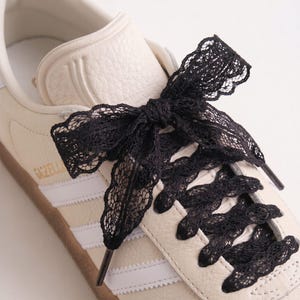 Puede incluir: Una zapatilla de color blanco roto con parte superior de cuero texturizado y tres rayas laterales blancas. El calzado cuenta con cordones de encaje negro atados en un lazo en la parte superior. La palabra GAZELLE está impresa en dorado.