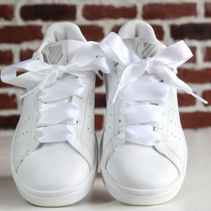 May include: A pair of white leather sneakers with white satin ribbon laces tied in bows. The sneakers have a classic design with a round toe and a white rubber sole.