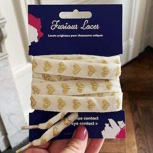 Op de afbeelding: Witte veters met gouden hartvormige accenten. De veters zijn verpakt op een blauwe kaart met de tekst "Furious Laces" en "Lacets originaux pour chaussures uniques".