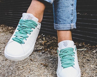 May include: White sneakers with green and white gingham laces. The shoes have a classic design with a low profile and a perforated upper. The laces are tied in a crisscross pattern.
