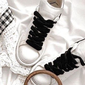 May include: A pair of white leather sneakers with black velvet laces. The laces are tied in a criss-cross pattern. The sneakers have a thick white sole and the text "Alexander McQueen" is printed on the side of one shoe.