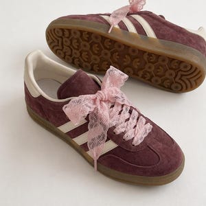 Op de afbeelding: Bordeaux suède sneakers met witte strepen en off-white accenten. De sneakers hebben roze kanten linten en een bruine zool. Eén schoen staat voor de andere, met de zool van de tweede schoen zichtbaar.