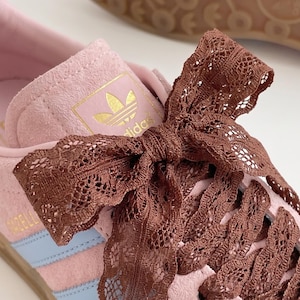 Può includere: Una sneaker in camoscio rosa chiaro con strisce laterali azzurre e un logo Adidas dorato sulla linguetta. La scarpa è allacciata con un ampio nastro di pizzo marrone scuro annodato in un grande fiocco. È visibile una suola in gomma sottostante.