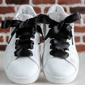 Può includere: Un paio di scarpe da ginnastica bianche con lacci in nastro di raso nero. Le scarpe hanno un design classico con un motivo traforato sui lati. I lacci neri sono annodati in grandi fiocchi.