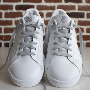 Può includere: Un paio di scarpe da ginnastica bianche in pelle con lacci grigi. Le scarpe hanno un design classico e sono perfette per l'uso quotidiano.