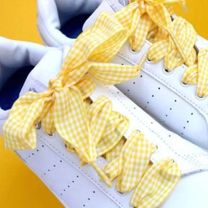 May include: White sneakers with yellow gingham ribbon laces tied in a bow. The laces are tied in a bow on the top of the shoe.