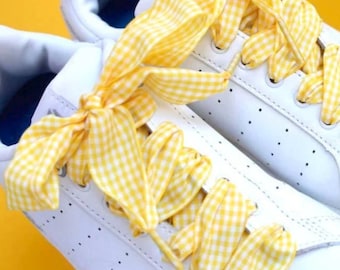 May include: White sneakers with yellow gingham ribbon laces tied in a bow. The laces are tied in a bow on the top of the shoe.