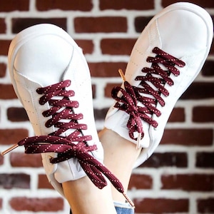 May include: White sneakers with burgundy laces featuring white polka dots. The laces are tied in a criss-cross pattern.