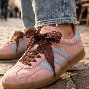 Op de afbeelding: Roze suède sneakers met lichtblauwe zijstrepen, een rubberen zool en donkerbruine veters van lint. De zijkant van de schoen toont de gouden tekst Gazelle en het Adidas-logo, met een lichtblauw detail op de hiel.