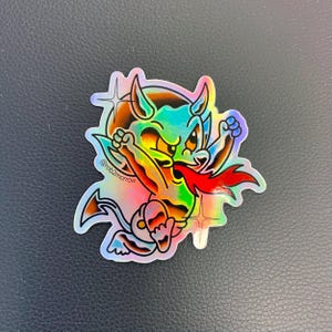 May include: A holographic sticker featuring a cartoon devil with red horns, a red tail, and a red tongue. The devil is wearing a green and blue outfit and has a mischievous expression. The sticker has a rainbow iridescent finish and includes the text "@rob2morrow".