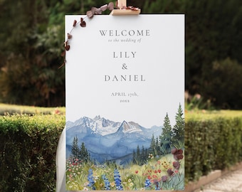 Könnte beinhalten: Ein weißes Hochzeits-Willkommensschild auf einer Holzstaffelei. Das Schild zeigt den Text "WELCOME to the wedding of LILY & DANIEL APRIL 17th, 20xx" über einem Aquarellgemälde von Bergen, Bäumen und Wildblumen.