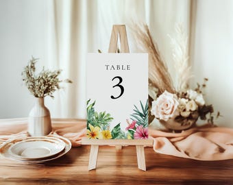 May include: A wooden easel displays a white sign with the word "TABLE" above the number "3". The sign is decorated with watercolor tropical flowers and foliage. A plate and vase are in the foreground.