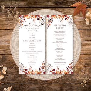 May include: Wedding ceremony programs with floral accents. The programs are white with text in a serif font. The programs are decorated with fall-colored leaves and flowers. The programs are on a woven placemat on a wooden surface.
