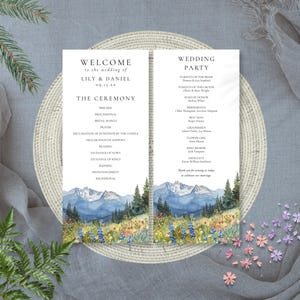 May include: Wedding ceremony program with a watercolor mountain landscape design. The program includes the names Lily & Daniel, the date, and the ceremony details. The program is set on a woven placemat with floral accents.