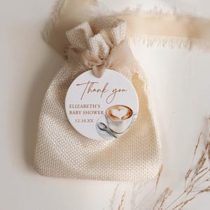 May include: A small, beige burlap bag tied with a cream-colored ribbon, featuring a round tag. The tag reads "Thank you" and "Elizabeth's Baby Shower 12.10.XX" with a watercolor image of a coffee cup with a heart-shaped design.