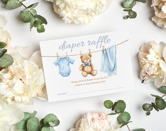 Op de afbeelding: Een baby shower luier verloting kaart met een aquarel illustratie van een blauwe romper, een teddybeer en een blauwe deken die aan een waslijn hangen. De kaart is omgeven door witte bloemen en groen. De tekst luidt "diaper raffle".