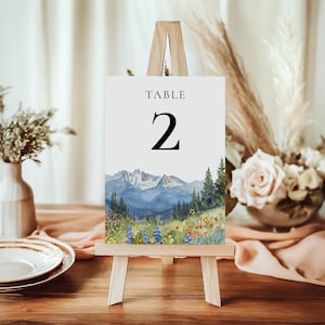 May include: A wooden easel displays a table number card with a watercolor mountain scene. The card reads "TABLE 2" in black text. The artwork features blue mountains, green trees, and colorful wildflowers. The scene is set on a wooden table.