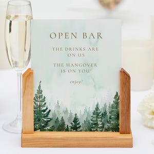 Op de afbeelding: Een houten open bar bord met de tekst "OPEN BAR" en "THE DRINKS ARE ON US THE HANGOVER IS ON YOU! enjoy!". Het bord heeft een aquarel bosontwerp en wordt in een houten standaard gehouden. Champagneglazen en een fles staan op de achtergrond.