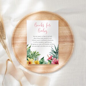 Puede incluir: Una tarjeta blanca con la inscripción rosa "Books for Baby", adornada con ilustraciones florales tropicales. La tarjeta está sobre una bandeja de madera, con espigas de trigo secas. El texto pide libros en lugar de tarjetas.