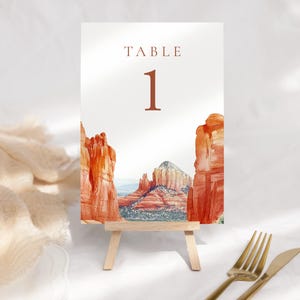 May include: A table number card with a watercolor landscape of red rock formations. The card reads "TABLE 1" in a brown serif font. The card is displayed on a small wooden easel, with gold-colored silverware in the foreground.