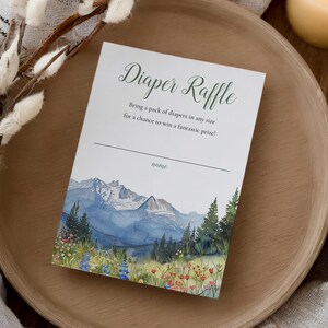 Puede incluir: Una tarjeta blanca con el texto "Diaper Raffle" en escritura verde. La tarjeta presenta una pintura de acuarela de una cordillera con tonos azules y verdes. La tarjeta está sobre una bandeja de madera.