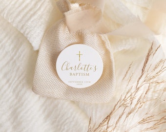 May include: A white round tag with gold lettering that says "Charlotte's Baptism" and the date "September 19th 20XX". A gold cross is above the name. The tag is sitting on a piece of beige fabric.