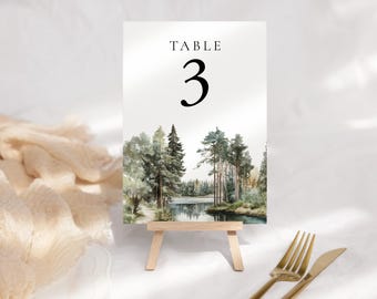 Op de afbeelding: Een tafelnummerkaart met het woord "TABLE" boven een groot zwart nummer 3. De kaart toont een aquarel schilderij van een boslandschap met bomen en een meer. De kaart wordt weergegeven op een kleine houten ezel, met goudkleurig bestek op de voorgrond.