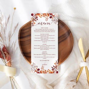 May include: A wedding menu card featuring a floral design in autumnal colours. The menu is on a wooden plate, with gold cutlery. The menu includes the names Elizabeth and James. The menu card is rectangular.