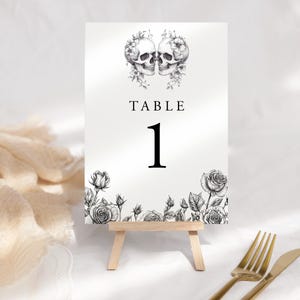 May include: A monochrome table number card featuring a floral and skull design. The card displays "TABLE 1" in a bold font. It's presented on a small wooden easel, with a gold fork and knife visible in the foreground.
