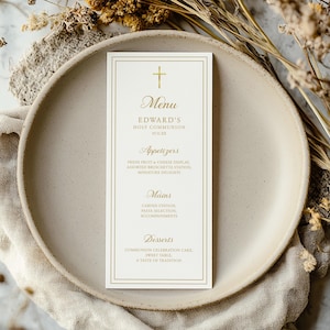May include: A cream-colored menu card with gold text and a cross at the top, displayed on a beige plate. The menu lists appetizers, mains, and desserts for Edward's Holy Communion. Dried flowers and a linen napkin surround the plate.