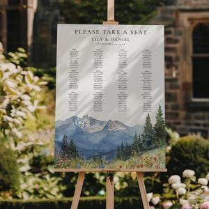 May include: A wedding seating chart on an easel. The sign reads "PLEASE TAKE A SEAT" above the names of Lily & Daniel and the date. The bottom of the sign features a watercolor painting of mountains, trees, and wildflowers.
